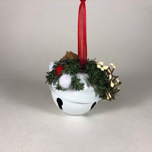 Op de afbeelding: Witte bel ornament met een rood lint en een groene en witte bloemstuk. De bel heeft een zwart geverfd ontwerp.