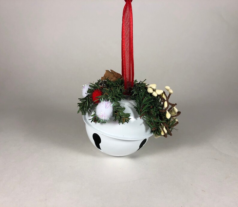 Op de afbeelding: Witte bel ornament met een rood lint en een groene en witte bloemstuk. De bel heeft een zwart geverfd ontwerp.
