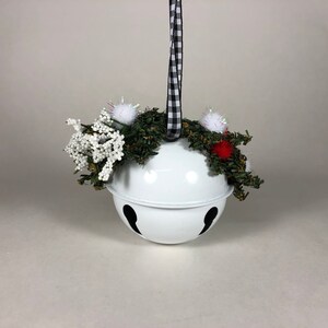 Op de afbeelding: Witte metalen jingle bell-versiering met een zwart-wit geblokt lint, versierd met een groene krans, witte en rode pompoenen en witte bessen takjes.