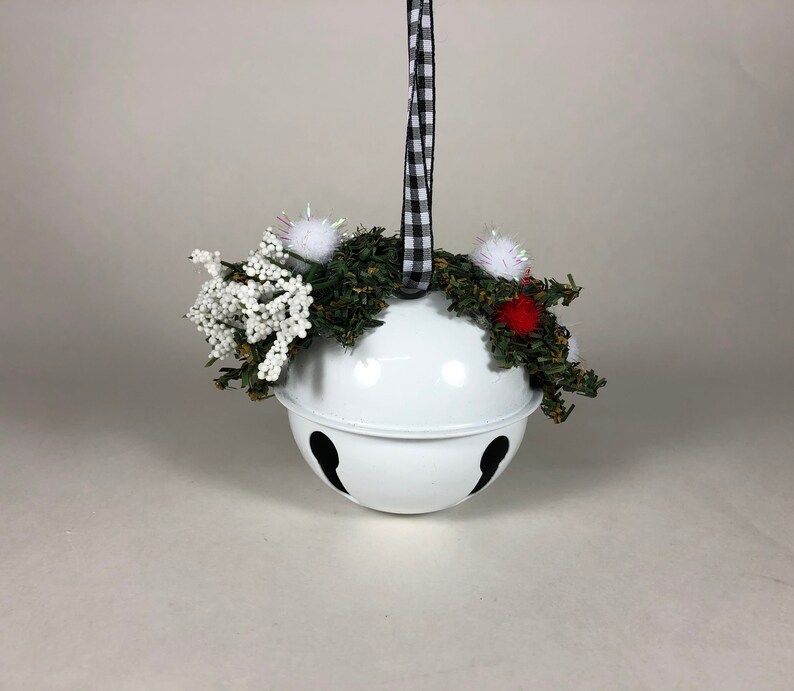 Op de afbeelding: Witte metalen jingle bell-versiering met een zwart-wit geblokt lint, versierd met een groene krans, witte en rode pompoenen en witte bessen takjes.
