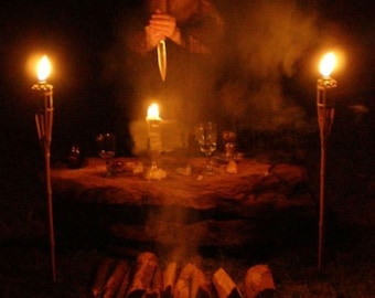 May include: A dimly lit scene with a person holding a knife in front of a table with candles, glasses, and a fire in the foreground. The person is wearing a dark hooded robe and is surrounded by smoke.