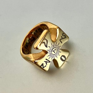 May include: Gold ring with a distinctive cross-shaped design. The ring features a sunburst motif with etched details and stylized symbols. The band has a smooth, polished finish, and the ring appears to be made of metal.