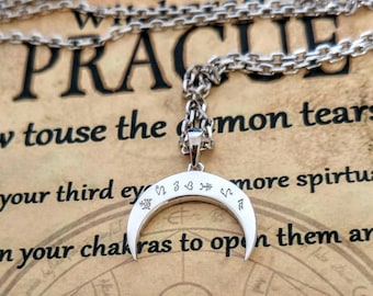 Puede incluir: Un collar con un colgante de media luna de plata, adornado con un pequeño diamante y símbolos grabados en la superficie. El collar está en una cadena de plata y descansa sobre un libro con la palabra "PRACTICE" visible.