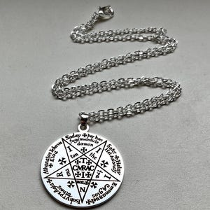 Peut inclure: Collier pendentif argenté. Le pendentif circulaire présente un design de pentagramme et du texte. Le pendentif est suspendu à une fine chaîne. Le pendentif comporte du texte sur le bord et au centre.
