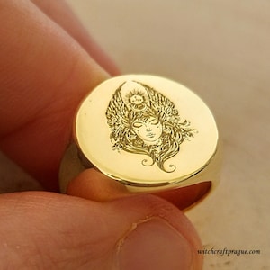 Könnte beinhalten: Ein goldener Siegelring mit einer gravierten Abbildung einer geflügelten Frau mit einer Krone. Die Frau hat die Augen geschlossen und ist von einem Heiligenschein umgeben. Der Ring ist ein Statement-Piece und wäre ein einzigartiges Geschenk.