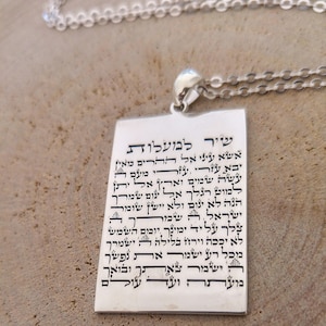 Könnte beinhalten: Eine silberne Halskette mit einem Anhänger mit hebräischem Text. Der Anhänger ist rechteckig und hat eine silberne Kette daran befestigt. Der Text auf dem Anhänger ist schwarz und ist ein Gebet oder ein Segen.