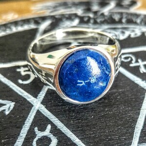 May include: A silver ring with a large, round, blue lapis lazuli stone set in the center. The ring is on a black surface with white markings.