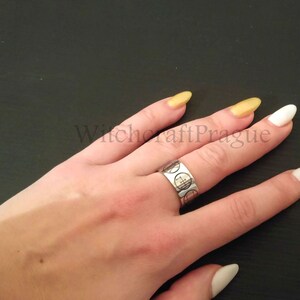Op de afbeelding: Een zilveren ring met een cirkelvormig ontwerp op een hand met witte en gouden nagellak. De ring heeft een herhalend patroon van cirkels met een kruis in het midden. De tekst "Witchcraft Prague" is zichtbaar op de achtergrond.