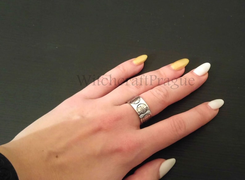 Op de afbeelding: Een zilveren ring met een cirkelvormig ontwerp op een hand met witte en gouden nagellak. De ring heeft een herhalend patroon van cirkels met een kruis in het midden. De tekst "Witchcraft Prague" is zichtbaar op de achtergrond.