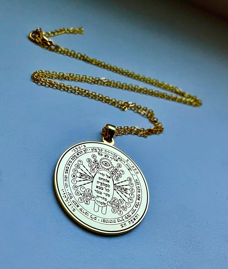 May include: Gold-colored pendant necklace with a circular charm. The charm features intricate engraved symbols and text, with a detailed design. The necklace chain is also gold-colored, and the pendant hangs from it. The background is a solid light blue.