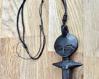 May include: A black wooden pendant necklace featuring a carved figure with a round head, facial features, and outstretched arms. The pendant is suspended from a black cord. The necklace is displayed on a wooden surface.