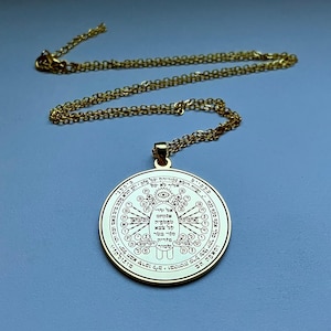 May include: A gold-colored pendant necklace with a circular charm. The charm features intricate engraved symbols and text, with a central eye design. The chain is also gold-colored, and the background is a solid light blue.