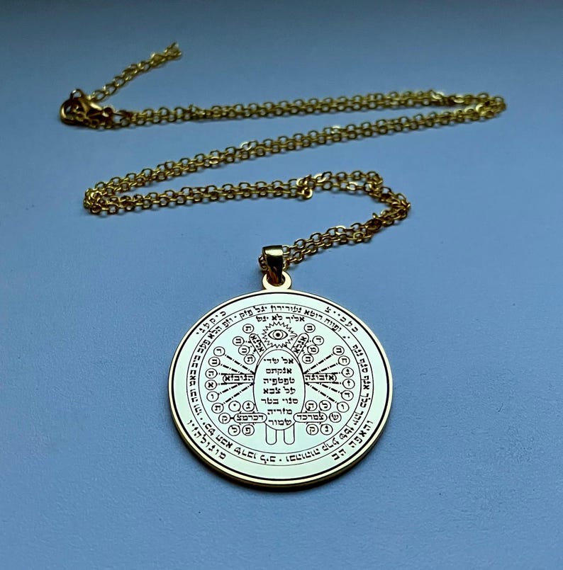 May include: A gold-colored pendant necklace with a circular charm. The charm features intricate engraved symbols and text, with a central eye design. The chain is also gold-colored, and the background is a solid light blue.