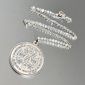 May include: A silver-colored pendant necklace with a circular charm. The charm features intricate Hebrew text and symbols around a central design. The chain is a delicate, linked style. The pendant is likely made of sterling silver or a similar metal.