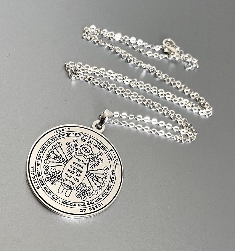 May include: A silver-colored pendant necklace with a circular charm. The charm features intricate Hebrew text and symbols around a central design. The chain is a delicate, linked style. The pendant is likely made of sterling silver or a similar metal.