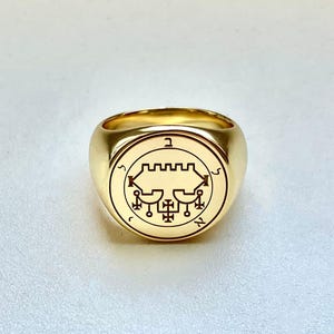 May include: A gold ring with a circular emblem featuring a detailed black design. The ring has a thick band and a polished finish. The emblem includes geometric shapes and symbols.