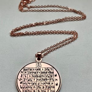 May include: A copper-coloured pendant necklace. The round pendant features an engraved grid pattern with Hebrew text and symbols. The necklace chain is also copper-coloured and delicate. The pendant has a loop for the chain to pass through.