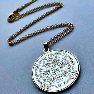 May include: A gold-colored pendant necklace with a circular charm. The charm features intricate engraved symbols and text, suspended from a delicate chain. The necklace is displayed on a light gray surface.