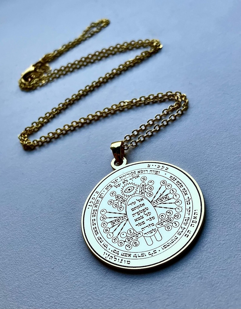 May include: A gold-colored pendant necklace with a circular charm. The charm features intricate engraved symbols and text, suspended from a delicate chain. The necklace is displayed on a light gray surface.