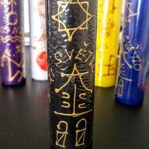 May include: A black cylindrical candle with a textured surface featuring gold hand-drawn symbols and the text SVETLO. The candle is positioned in front of other colored candles including purple, white, yellow, and blue, each with similar gold markings.