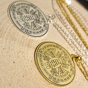 May include: Two circular pendants with intricate designs and text, one silver and one gold, each suspended on a delicate chain. The pendants rest on a light-colored, textured surface. The website address "witchcraftprague.com" is visible at the bottom.