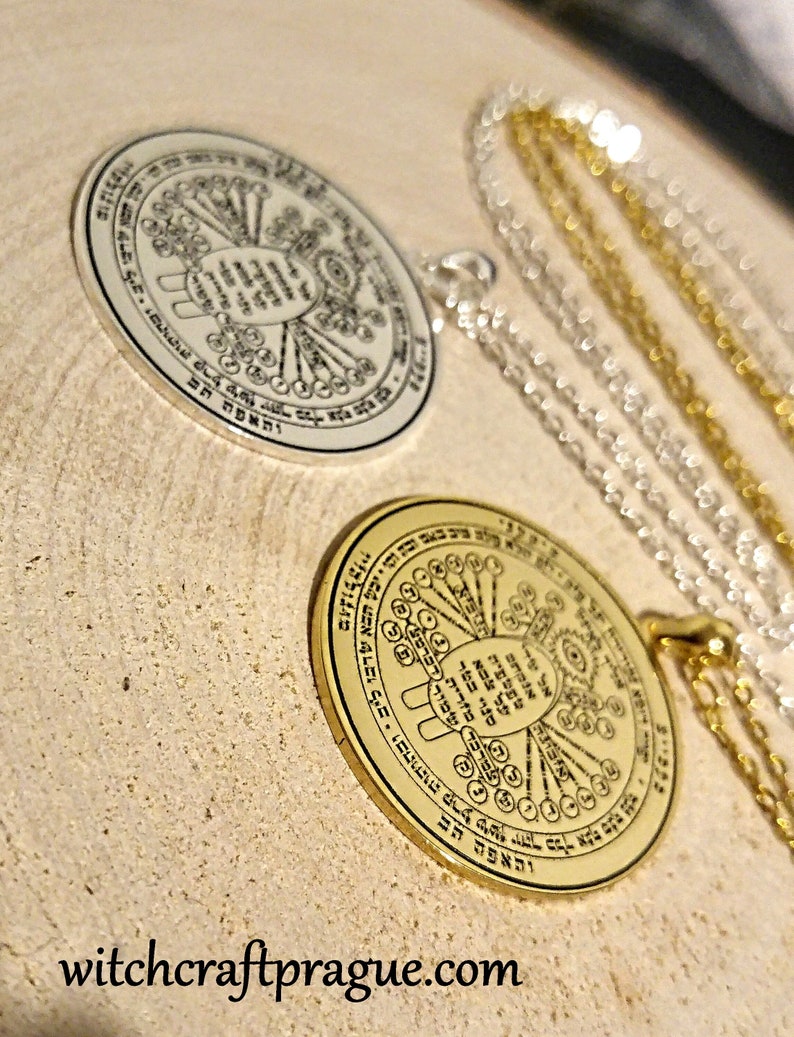 May include: Two circular pendants with intricate designs and text, one silver and one gold, each suspended on a delicate chain. The pendants rest on a light-colored, textured surface. The website address "witchcraftprague.com" is visible at the bottom.