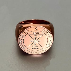Puede incluir: Un anillo de color cobre con una cara circular con texto hebreo y diseños geométricos. El anillo tiene un acabado pulido y una banda ancha. El texto incluye las palabras "Adonai" y "Elohim".