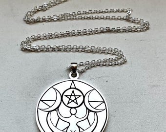 Peut inclure: Collier pendentif argenté avec un pendentif circulaire. Le pendentif présente un motif détaillé avec une étoile centrale, des croissants de lune et une figure féminine stylisée. La chaîne est délicate et argentée. Bijou.