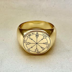 Può includere: Un anello sigillo in oro lucido con una faccia circolare incisa con un complesso sigillo geometrico. Il design include un motivo centrale a forma di stella con linee radianti, piccoli accenti circolari e caratteri ebraici lungo il bordo.