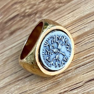 May include: A gold ring with a textured band and a circular, grey-toned centerpiece. The centerpiece features an intricate design with a tree or plant-like motif. The ring is set against a wooden surface.