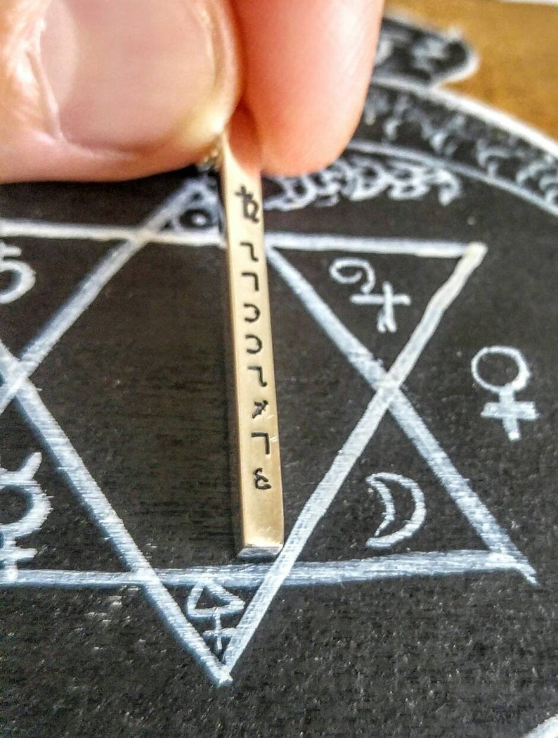 May include: A silver bar pendant with Hebrew text inscribed on it. The pendant is resting on a black surface with a white star of David drawn on it.