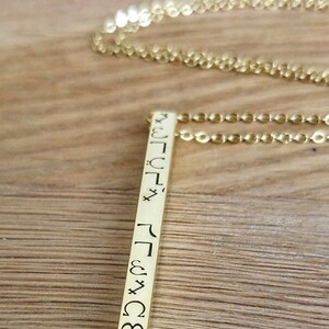 May include: Gold necklace with a long, thin pendant featuring a vertical row of symbols. The pendant is engraved with a series of symbols, possibly runes or other ancient characters.