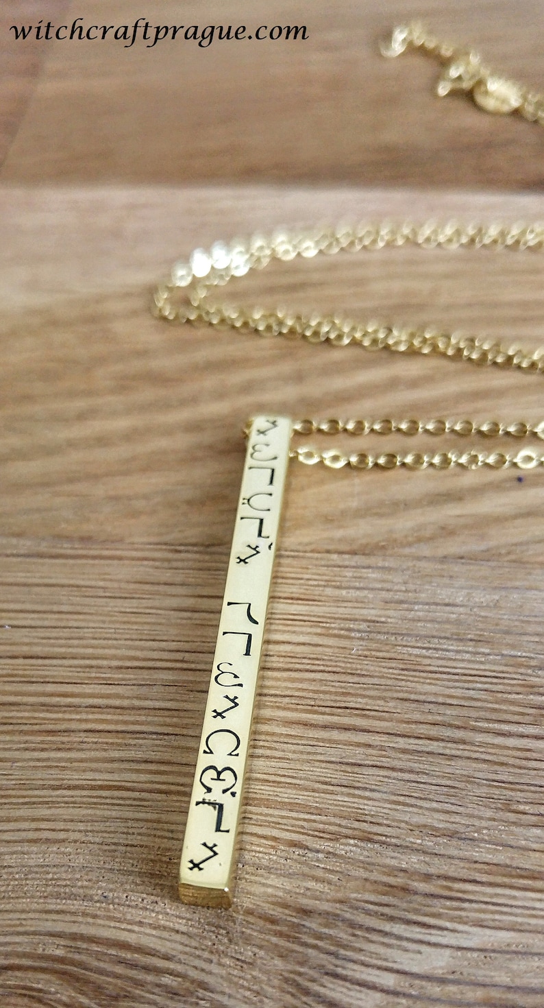 May include: Gold necklace with a long, thin pendant featuring a vertical row of symbols. The pendant is engraved with a series of symbols, possibly runes or other ancient characters.