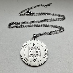 Op de afbeelding: Een zilverkleurige ronde hanger aan een bijpassende metalen ketting met een karabijnsluiting. De hanger is voorzien van een centraal vijf-bij-vijf raster met cijfers, Hebreeuwse tekens en het symbool van de planeet Mars.