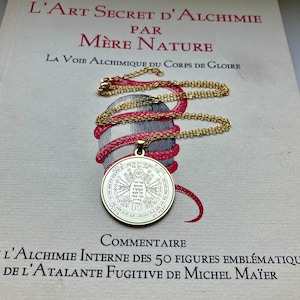 May include: A gold-colored pendant necklace with a detailed circular design, resting on a book with French text. The pendant hangs from a delicate chain. The book's title is "L'Art Secret D'Alchimie". The necklace is a symbol of spirituality and mysticism.