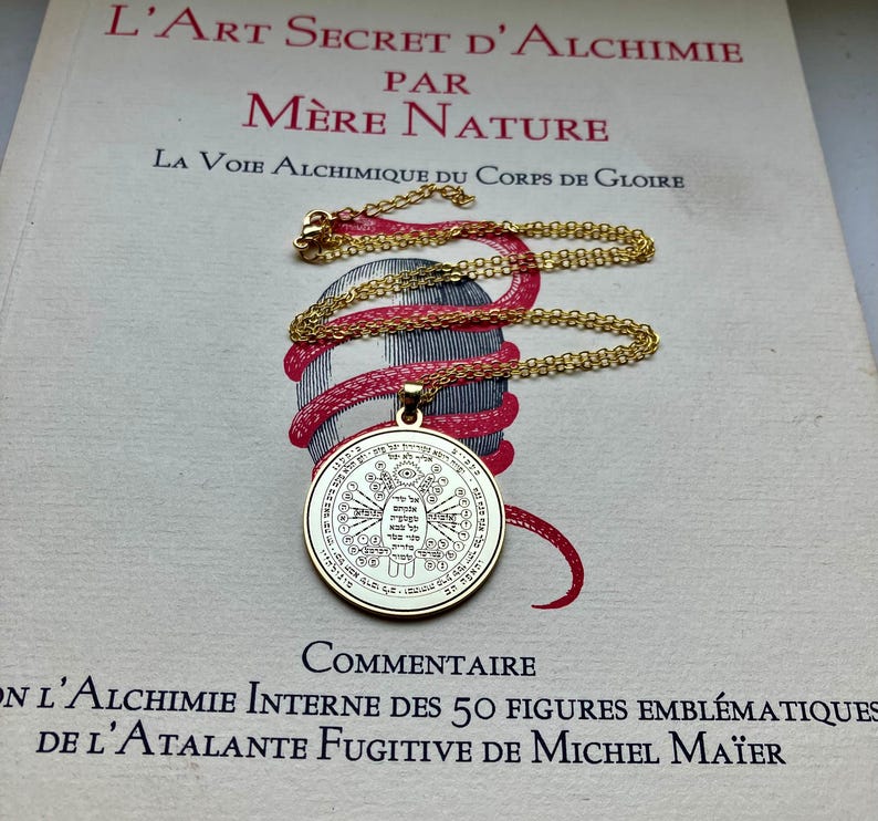 May include: A gold-colored pendant necklace with a detailed circular design, resting on a book with French text. The pendant hangs from a delicate chain. The book's title is "L'Art Secret D'Alchimie". The necklace is a symbol of spirituality and mysticism.