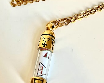 Könnte beinhalten: Goldfarbene Halskette mit zylindrischem Anhänger. Der Anhänger hat ein klares Glasröhrchen mit weißem Papier und roten und schwarzen Designs. Goldfarbene Kappen mit schwarzen Symbolen verzieren den Anhänger. Die Kette ist ebenfalls goldfarben.