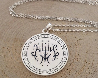 Op de afbeelding: Zilveren ketting met een ronde hanger met een zwart-wit symbool met een drietand en andere markeringen. De hanger is omgeven door een cirkel met tekst in een vreemde taal.