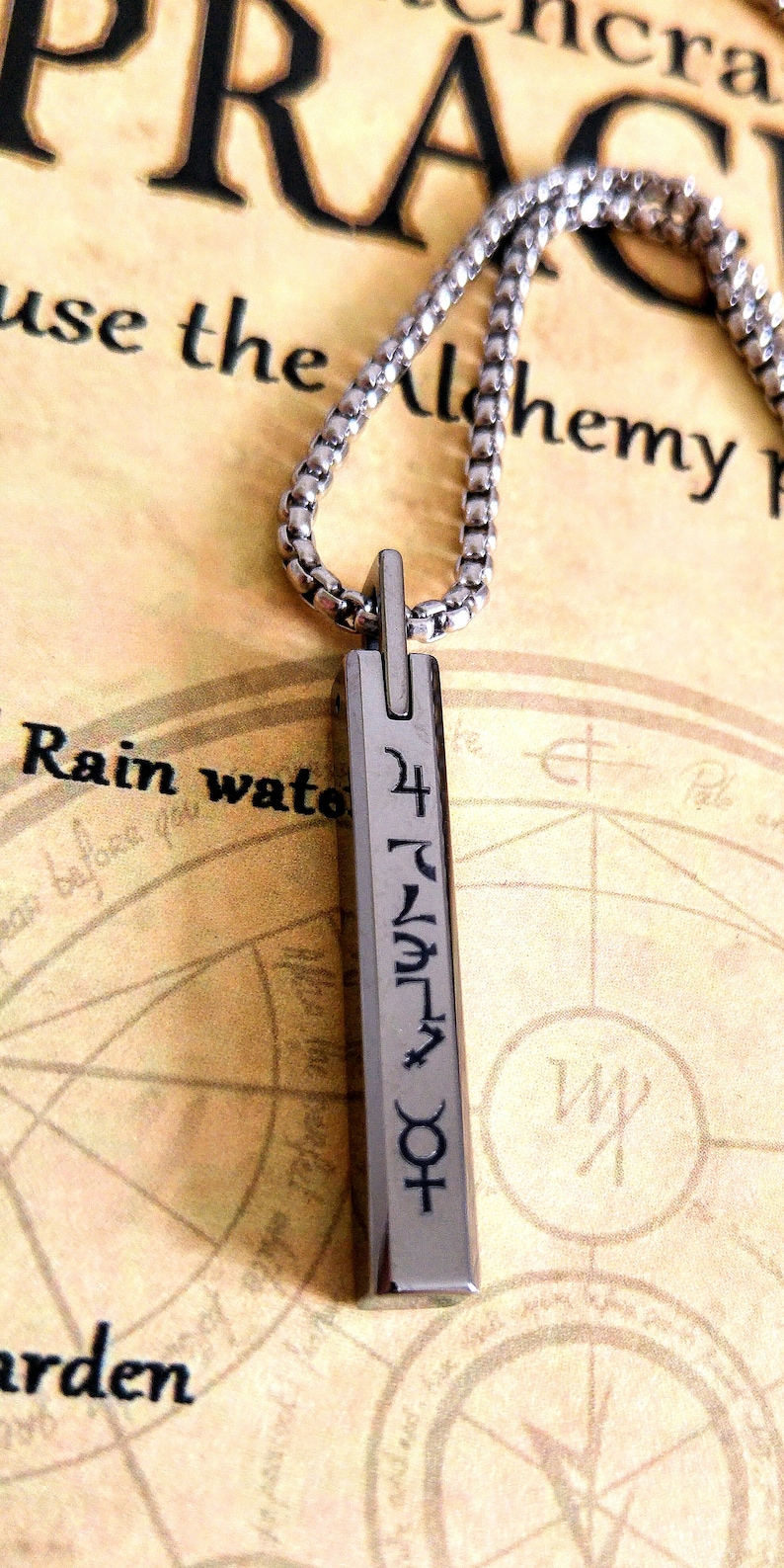 May include: A silver chain necklace with a silver pendant. The pendant is rectangular and has a symbol on it. The symbol is a combination of letters and symbols. The necklace is lying on a piece of paper with a map on it.