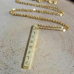 May include: A gold chain necklace with a vertical bar pendant. The bar has various symbols engraved on it, including a crescent moon, a heart, a star, and a sun.