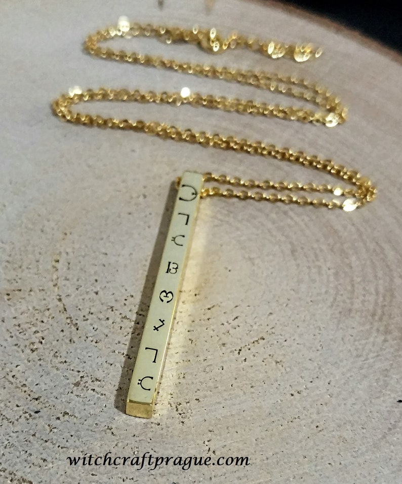 May include: A gold chain necklace with a vertical bar pendant. The bar has various symbols engraved on it, including a crescent moon, a heart, a star, and a sun.