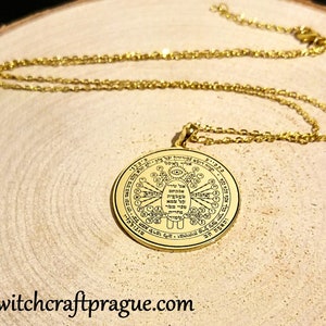 May include: Gold-colored pendant necklace with a detailed circular design and chain. The pendant features intricate symbols and text. The necklace is displayed on a wooden surface. The website address "witchcraftprague.com" is visible at the bottom.
