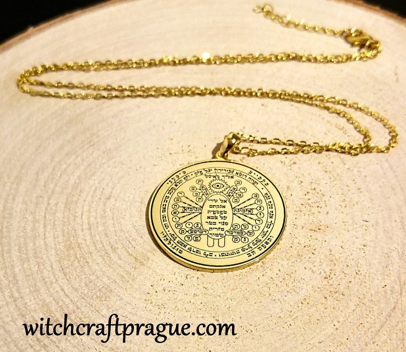 May include: Gold-colored pendant necklace with a detailed circular design and chain. The pendant features intricate symbols and text. The necklace is displayed on a wooden surface. The website address "witchcraftprague.com" is visible at the bottom.