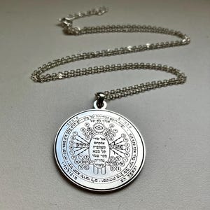 May include: A silver-colored pendant necklace. The pendant is round with intricate engraved designs and text. The necklace chain is also silver. The pendant features a central eye symbol and other symbols and text around it.