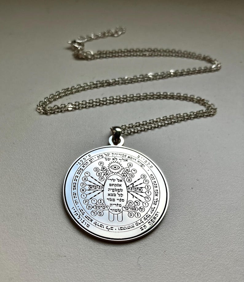 May include: A silver-colored pendant necklace. The pendant is round with intricate engraved designs and text. The necklace chain is also silver. The pendant features a central eye symbol and other symbols and text around it.