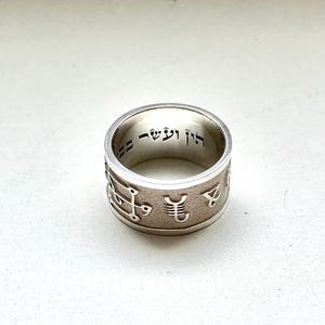 May include: A silver ring with etched symbols and Hebrew text inside. The ring has a wide band with a brushed metal finish. The symbols are evenly spaced around the band. The Hebrew text is in a simple font.