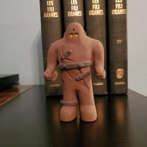 Könnte beinhalten: Eine tonfarbene, humanoide Figur mit einem glatten, abgerundeten Kopf, zwei leuchtend gelben Augen und einem kleinen Punkt auf der Stirn. Die Figur hat dunkelbraune Riemen um den Oberkörper und ein Bein gewickelt und eine matte Oberfläche.