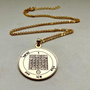 Puede incluir: Collar con colgante dorado con un colgante circular. El colgante presenta una cuadrícula de números y texto hebreo. La cadena y el colgante son de color dorado brillante. El colgante tiene el número 999.