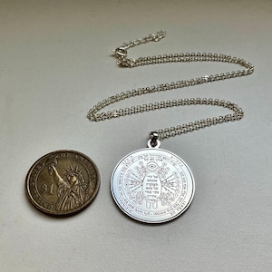 May include: A silver pendant necklace with a circular charm featuring intricate engraved symbols and text. The necklace chain is also silver. A US one-dollar coin is next to the pendant for size comparison.