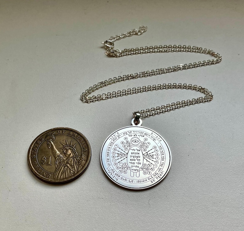 May include: A silver pendant necklace with a circular charm featuring intricate engraved symbols and text. The necklace chain is also silver. A US one-dollar coin is next to the pendant for size comparison.